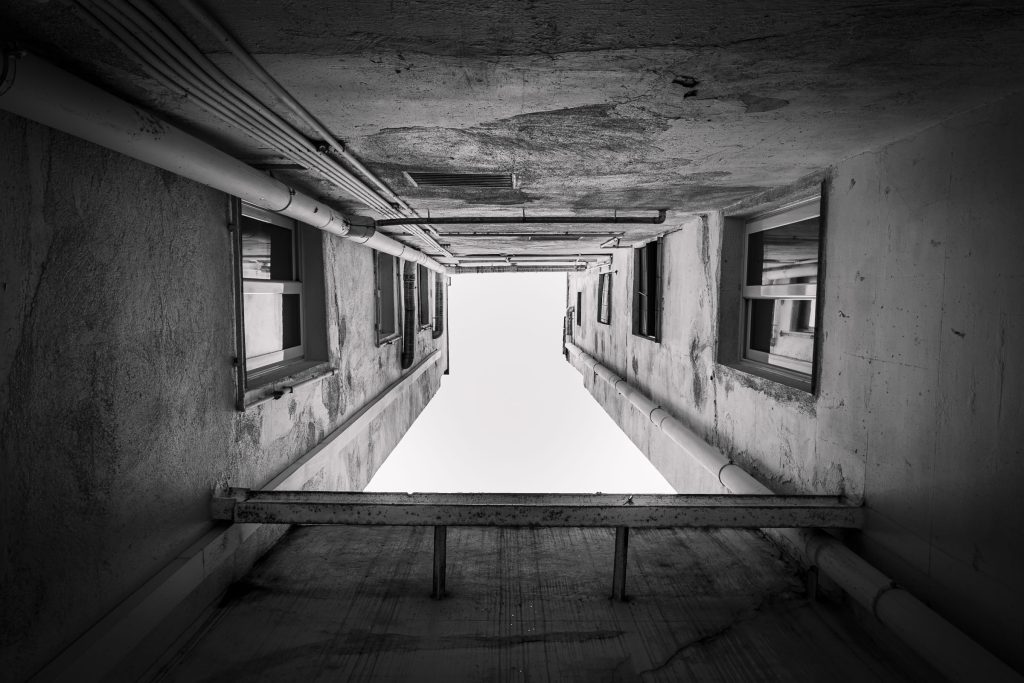 Black and white architectural photograph looking up through a narrow concrete courtyard with symmetrical walls, windows and exposed pipework.