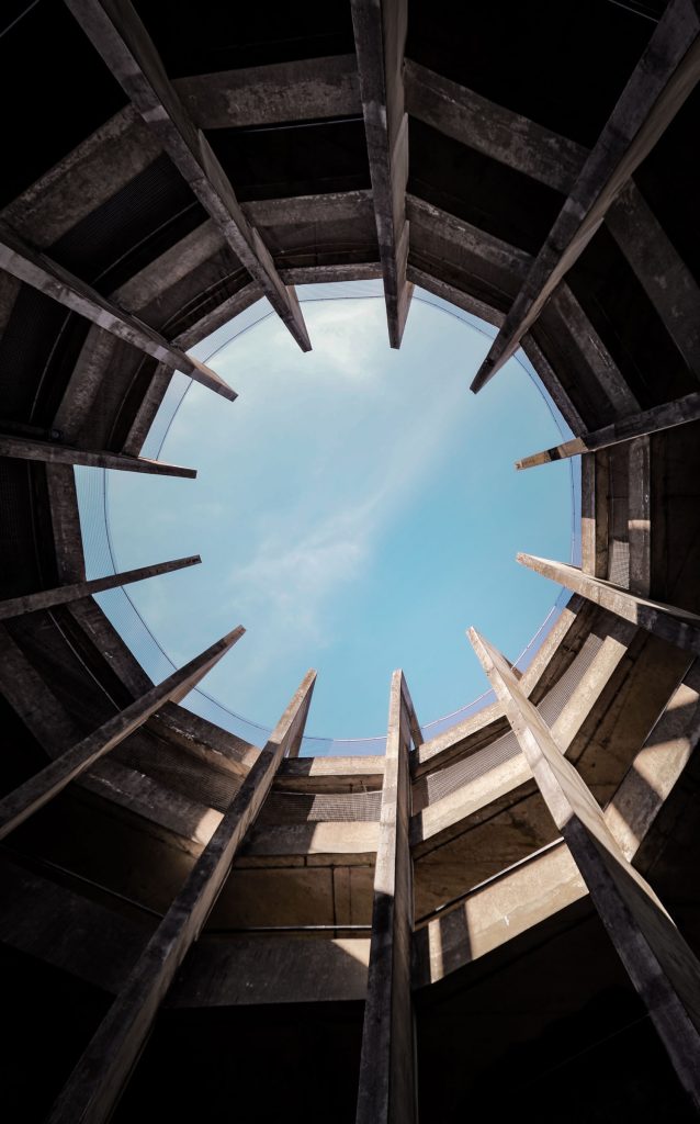 Upward architectural photograph of a circular concrete structure with radial beams framing a blue sky.