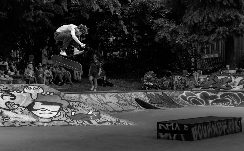 Black and white action photograph of a skateboarder performing a kickflip over a bowl while spectators watch.