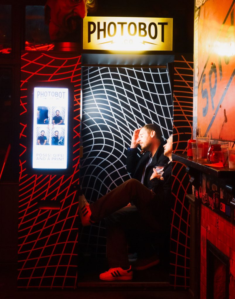 Cinematic nightlife photograph of two people sitting inside a brightly lit photobooth surrounded by neon lights.