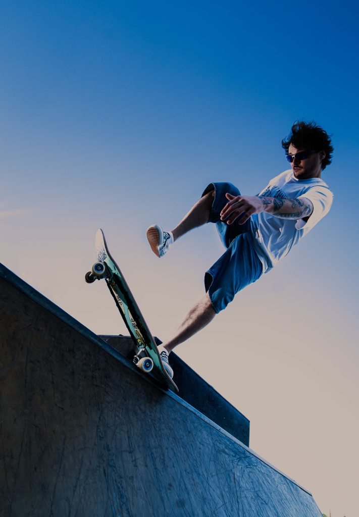 Low-angle photograph of a skateboarder balancing on the edge of a concrete skate ramp against a clear blue sky.