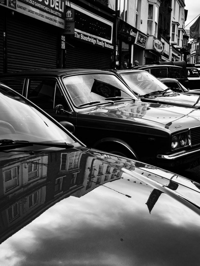 Black and white photograph of classic cars parked in town with reflections across polished bodywork.