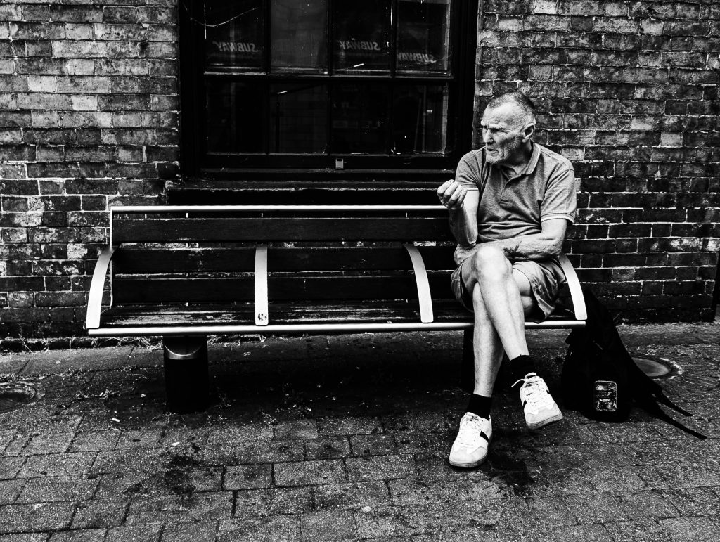 Black and white street portrait of an elderly man sitting on a bench outside a brick building.